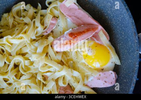 La pasta Fettuccine con uova fritte e salsicce è cucinata in una padella condita con pepe nero macinato ed erbe aromatiche, per un pasto semplice e veloce Foto Stock