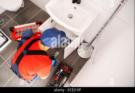 Un idraulico esperto ispeziona e ripara un armadietto del lavandino del bagno, utilizzando vari strumenti e attrezzature in una casa contemporanea durante le ore diurne. Foto Stock