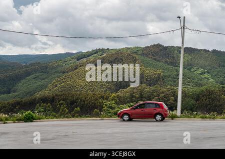 Auto compatta Red Ford Fiesta parcheggiata su un punto panoramico pavimentato con vista panoramica su lussureggianti montagne e foreste verdi sotto un cielo nuvoloso. Foto Stock