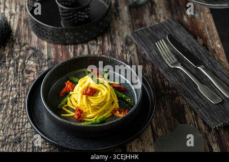 Spaghetti gourmet con asparagi e pomodori secchi serviti su un elegante piatto nero su un rustico tavolo in legno Foto Stock