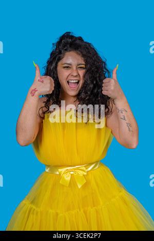 Giovane donna eccitata che indossa un abito giallo che mostra i pollici in alto, celebrando il successo e l'approvazione su sfondo blu Foto Stock
