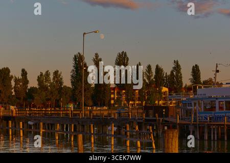 Vista dal molo verso gli hotel illuminati sul lago sotto il vivace tramonto dorato. La luna splende sopra, l'acqua rispecchia la luce, aggiungendo un tocco romantico al veicolo elettrico Foto Stock