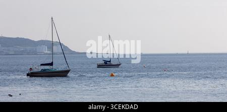 Due yacht ancorati su calme acque marine con edifici industriali e verdi colline sullo sfondo, scenario costiero in una giornata nebbiosa. Foto Stock