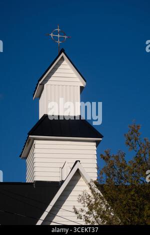 primo piano della parte superiore del campanile bianco della chiesa con croce e cerchio sul tetto nero superiore con alberi in basso a destra cielo blu profondo nella stanza dello sfondo Foto Stock