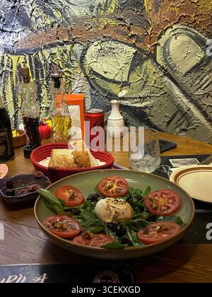 Questa immagine mostra un'insalata di burrata fresca con fette di pomodoro, noci, olive e verdure a foglia verde. Pane in cesto rosso, olio d'oliva, aceto, Foto Stock