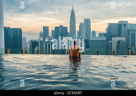 Uomo turista che si rilassa nella piscina a sfioro sul tetto all'alba, Kuala Lumpur, Malesia. Viaggiatore di sesso maschile che gode di vista sulla città con servizi finanziari del centro Foto Stock