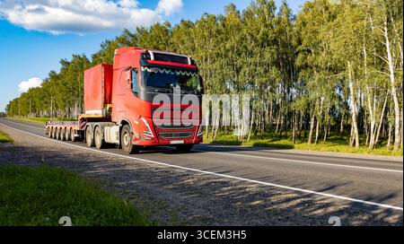 Grande camion rosso con rimorchio a pianale per attrezzature pesanti su una strada di campagna in movimento contro il cielo blu e la foresta. Foto Stock