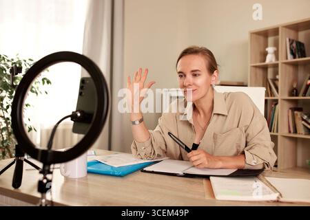 Una donna è seduta a una scrivania in un ufficio domestico, impegnata in una riunione online con uno smartphone fissato su un supporto mobile e illuminato da un anello Foto Stock