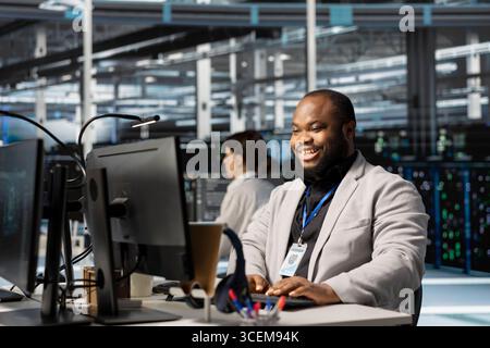 Felice specialista IT di hub server entusiasta dopo aver completato con successo le operazioni. Felice uomo afroamericano nel data center che controlla lo stato dell'infrastruttura rapporto che mostra indicatori di prestazioni stabili Foto Stock