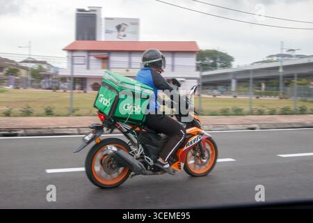 Corriere motociclistico che consegna il cibo in bicicletta lungo una strada urbana con edifici e vegetazione sullo sfondo George Town Penang Island malesia Foto Stock