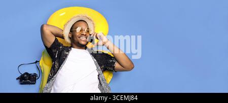 Funky ragazzo nero in abiti estivi alla moda sdraiato su gonfiabile lilo, parlando su smartphone su sfondo blu, vista dall'alto Foto Stock