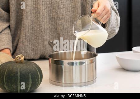 Una donna versa il latte in una pentola in una cucina moderna, cucinando piatti di zucca. Ricette autunnali Foto Stock