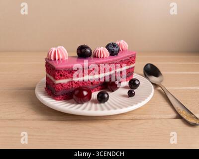 Appetitosa fetta di torta rosa con ripieno di panna, ricoperta di mirtilli e ciliegie, servita su un piatto bianco con un cucchiaio. Il dessert perfetto ph Foto Stock