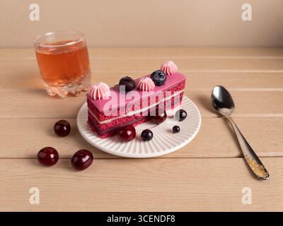 Una fetta di torta a strati con glassa rosa, adornata con mirtilli e ciliegie, accompagnata da una bevanda color arancio e da un cucchiaio decorativo, Foto Stock