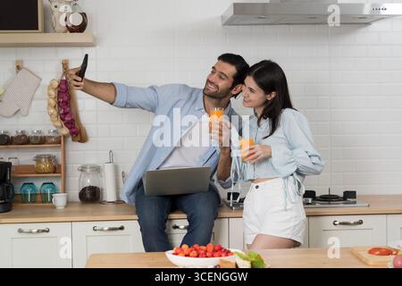 Giovane coppia in una cucina moderna sorridente mentre scatta selfie con lo smartphone, tenendo bicchieri di succo d'arancia, felicità, stile di vita, relazione, casua Foto Stock