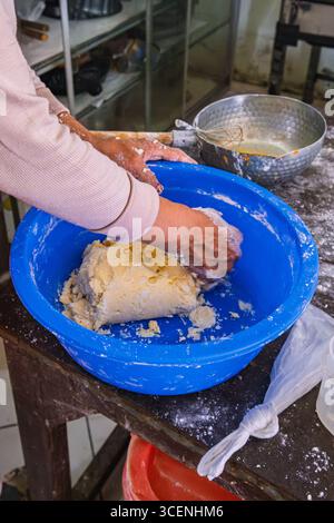 Primo piano delle mani impastando l'impasto in un recipiente blu durante la preparazione del pane in una cucina da forno. Foto Stock