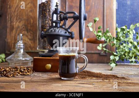 Tazza di caffè nero appena preparato su un tavolo in legno rustico con caffè macinato, chicchi e un macinacaffè vintage sullo sfondo, in un ambiente accogliente Foto Stock