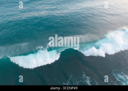 Vista aerea di un'onda ondulata che si insinua e si schianta sulle rocce scure sottostanti, mostrando le contrastanti acque turchesi e gli spruzzi bianchi schiumosi, Uluwatu, Bali, Indonesia. Foto Stock