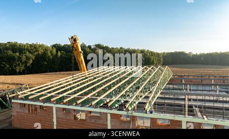 Un grande cantiere con una struttura del tetto parzialmente costruita in acciaio verde e una gru gialla visibile, adagiata su uno sfondo rurale Foto Stock