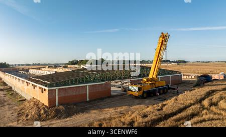 Una gru telescopica mobile che solleva il tetto in acciaio verde fa leva su una grande struttura in mattoni in un cantiere rurale, circondato da campi di grano e c Foto Stock