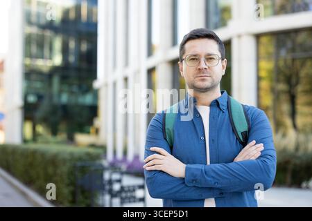 Un giovane uomo che indossa occhiali e uno zaino posa con sicurezza davanti a un edificio del campus. L'ambiente cattura un'atmosfera educativa moderna e urbana Foto Stock