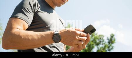 Primo piano di mans sportivi con smartwatch che regge il telefono cellulare. Controlla le app, concentrati sul dispositivo. Avanzamento fitness su app. Banner. Foto Stock