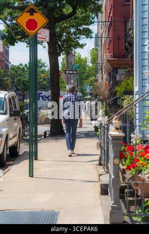 Brooklyn, Stati Uniti - 13 agosto 2025: Vista di un pedonale che passeggia lungo un marciapiede soleggiato fiancheggiato da pietre arenarie e vegetazione lussureggiante. Foto Stock