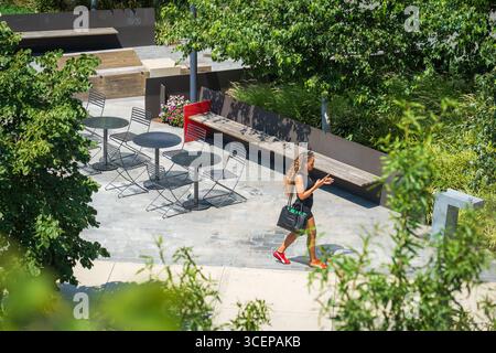 Brooklyn, Stati Uniti - 13 agosto 2025: Vista di una donna che cammina attraverso un moderno spazio parco con tavoli, sedie e panchine, incorniciato da una vegetazione lussureggiante. Foto Stock