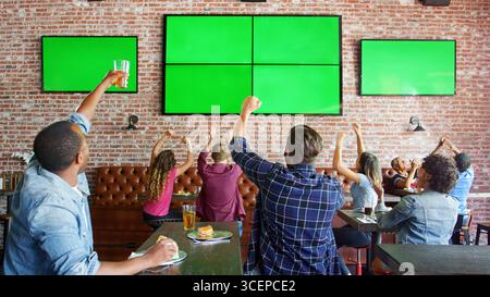 Le persone sedute ai tavoli del bar dello sport guardano una partita sui green screen e festeggiano Foto Stock