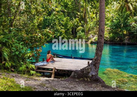 Donna in costume da bagno rosso seduta su un molo di legno vicino a un lago turchese nella natura tropicale. Foto Stock
