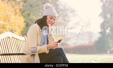 Donna a piedi seduta sulla panchina nel parco autunnale o in campagna che controlla il telefono cellulare Foto Stock