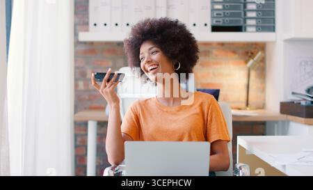 Giovane donna d'affari sorridente che lavora alla scrivania in ufficio e parla con il microfono del telefono cellulare Foto Stock