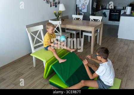 Due fratelli che giocano con la palla su scivolo improvvisato fatto di tappetini da palestra nella cucina moderna di casa il giorno d'estate. Concetto di attività creative al chiuso Foto Stock