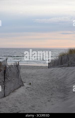 Un sentiero che attraversa una recinzione in listelli di legno consumati e porta a un sentiero sabbioso sulla spiaggia verso acque oceaniche calme durante un dolce tramonto Foto Stock