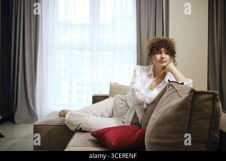 La donna si rilassa sul divano in un elegante soggiorno durante il pomeriggio soleggiato a casa Foto Stock