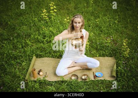 Una persona siede a gambe incrociate su un tappeto circondato da ciotole cantanti, impegnandosi in una pratica di meditazione. L'erba verde e la natura creano una tranquilla atmos Foto Stock