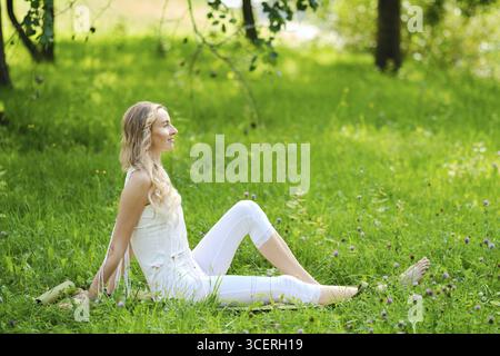 Una giovane donna con i capelli lunghi siede serenamente sull'erba in un vibrante prato verde. Indossa un vestito leggero e si rilassa pacificamente, circondato da un soffio Foto Stock