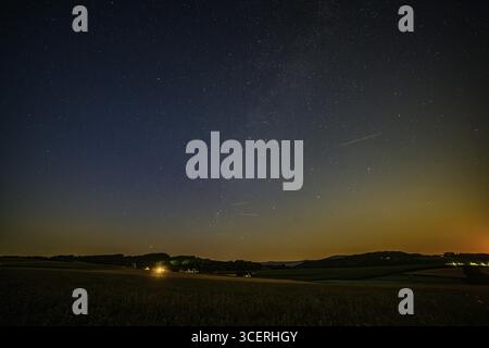 Sotto il cielo sereno, numerose stelle brillano sopra una tranquilla scena rurale della bassa Baviera. Una stella cadente dei Perseidi si muove attraverso il cielo verso Foto Stock