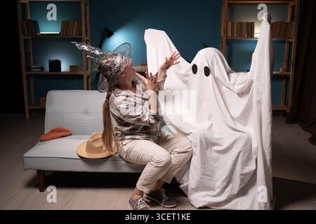 Amici che celebrano Halloween a casa, uno in costume da fantasma e una donna in cappello da strega Foto Stock