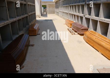 Le bare dei migranti morti nel naufragio del 13 agosto 2025 riposano nel nuovo cimitero di Cala Pisana, Lampedusa. Alcuni recano lettere, probabilmente per l'identificazione. Nella camera mortuaria, le persone si riuniscono in preghiera, mentre i fiori giacciono alla porta. Un tavolo di chirurgo utilizzato per le autopsie si trova nelle vicinanze, sottolineando la tragedia. Le vittime saranno poi trasferite nei cimiteri in Sicilia. Foto Stock