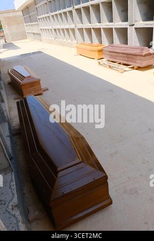 Le bare dei migranti morti nel naufragio del 13 agosto 2025 riposano nel nuovo cimitero di Cala Pisana, Lampedusa. Alcuni recano lettere, probabilmente per l'identificazione. Nella camera mortuaria, le persone si riuniscono in preghiera, mentre i fiori giacciono alla porta. Un tavolo di chirurgo utilizzato per le autopsie si trova nelle vicinanze, sottolineando la tragedia. Le vittime saranno poi trasferite nei cimiteri in Sicilia. Foto Stock