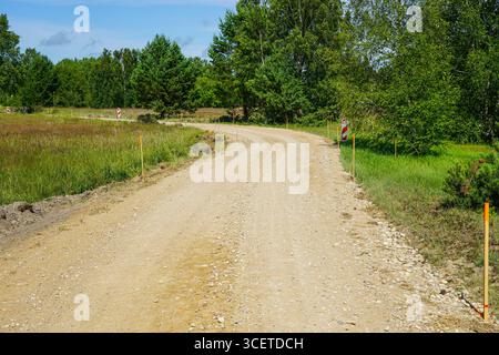 Una tortuosa strada di ghiaia in costruzione segnata da picchetti e segnali di avvertimento, circondata da campi verdi e alberi estivi, Lettonia, Europa Foto Stock