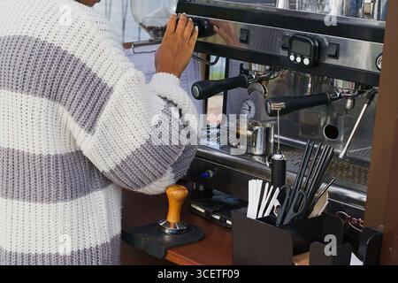 Barista con pelle scura in maglione a righe per preparare l'espresso in una macchina da caffè professionale. Primo piano di mani e attrezzature in fase di lavoro con wa Foto Stock