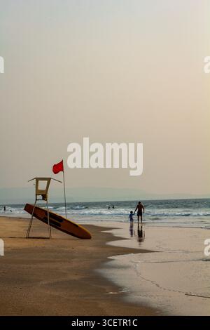 Questa immagine mostra una torre bagnino con una bandiera rossa e una tavola da surf di salvataggio su una spiaggia sabbiosa, con poche persone e dolci onde sullo sfondo Foto Stock