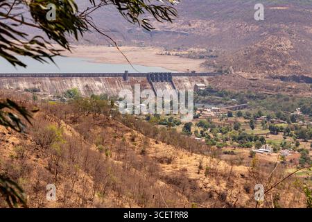 Questa immagine mostra una grande diga annidata in un paesaggio collinare asciutto, che si affaccia su una valle verde con case e alberi sparsi, Foto Stock