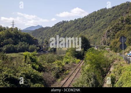 paesaggio montano con un fiume che scorre attraverso una lussureggiante foresta, un bellissimo ponte ad arco che attraversa l'acqua e binari ferroviari che corrono lungo il ban Foto Stock