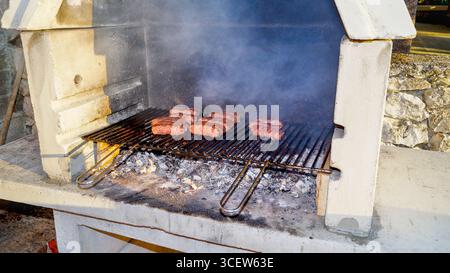 Grigliate Cevapcici su un barbecue rustico in mattoni all'aperto: Tradizionali salsicce balcaniche cucinate sul fuoco aperto in giardino Foto Stock