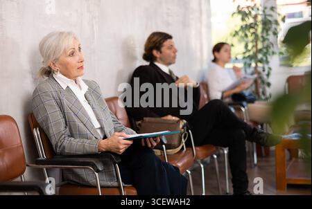 Una donna anziana e sicura di sé è seduta nella hall di una grande azienda, in attesa del suo turno per vedere uno specialista in ufficio Foto Stock
