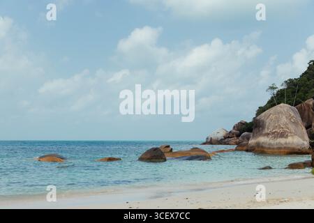 Le limpide acque turchesi si infrangono dolcemente sulla spiaggia sabbiosa, con grandi rocce sparse lungo la spiaggia e lussureggiante vegetazione sullo sfondo sotto un br Foto Stock