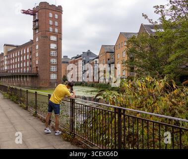 Un uomo appoggiato su una ringhiera e che scatta una foto lungo un passaggio pedonale lungo il fiume, con edifici storici in mattoni e appartamenti moderni sullo sfondo. Urbano Foto Stock